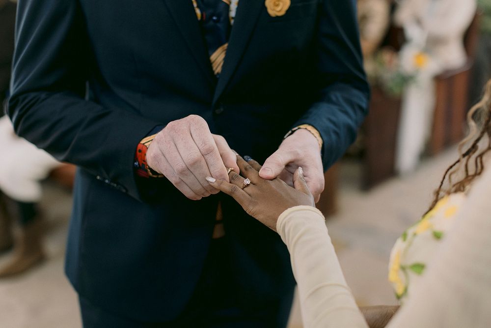 Groom placing a wedding ring on bride's finger during a ceremony, wearing a navy suit with a patterned tie.