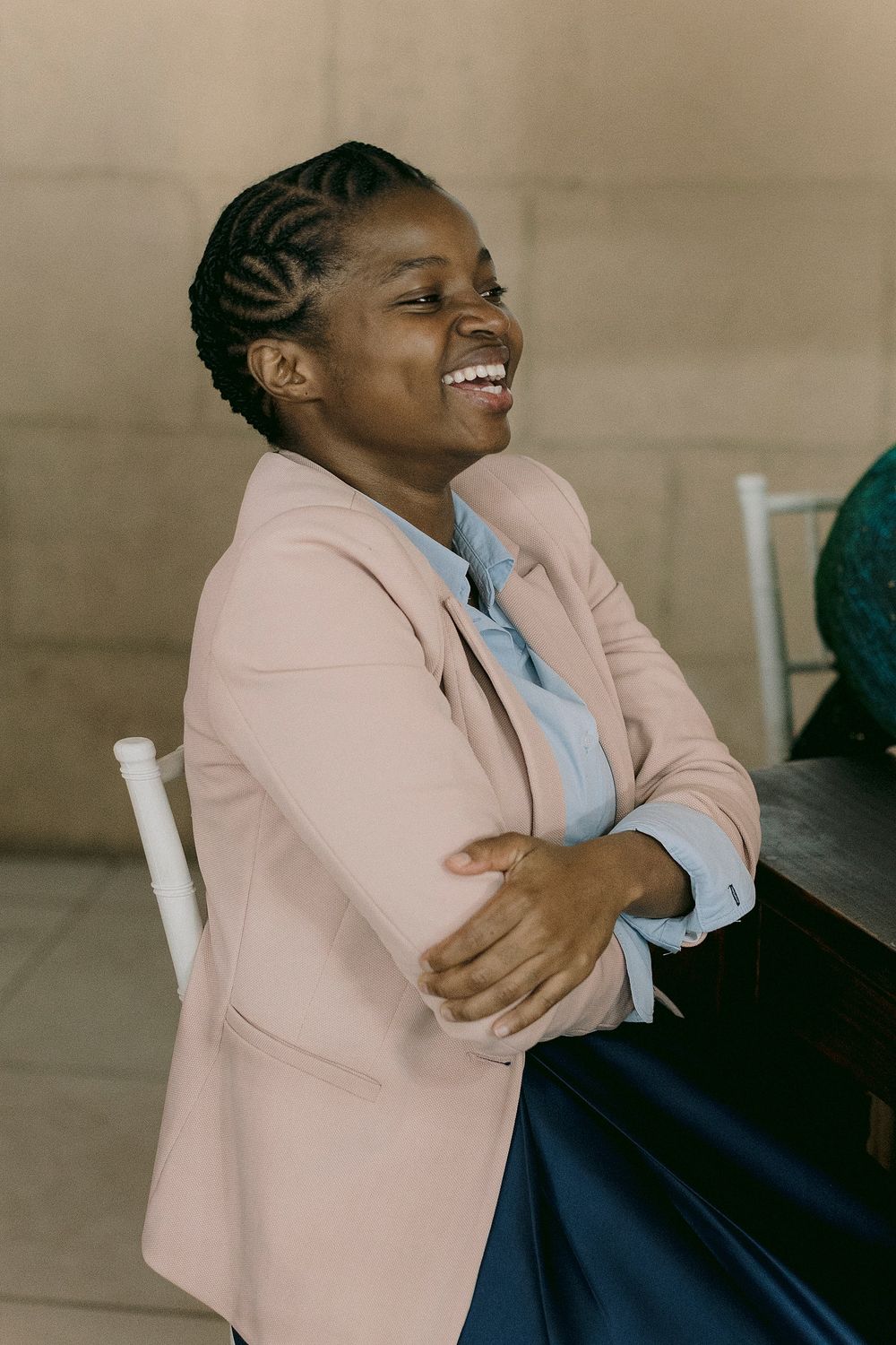 A joyful Black woman in a pink blazer laughs while seated, wearing braided hair and a light blue shirt.