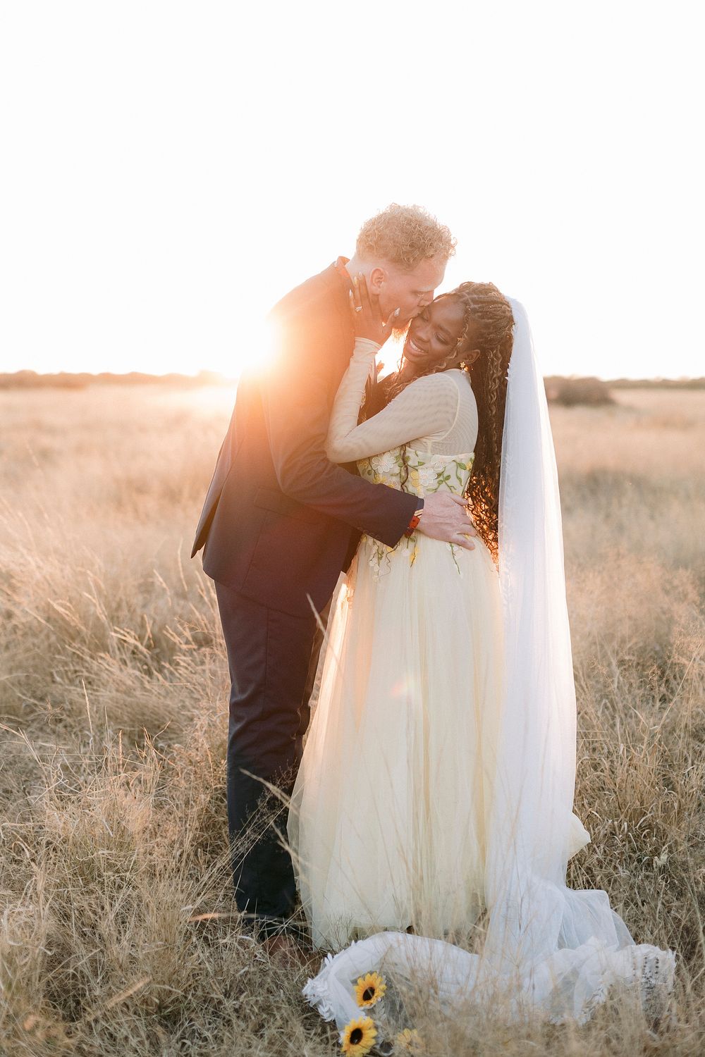 A couple embraces in a golden field at sunset, bride in white gown with veil, groom in dark suit, sunflowers at their feet.