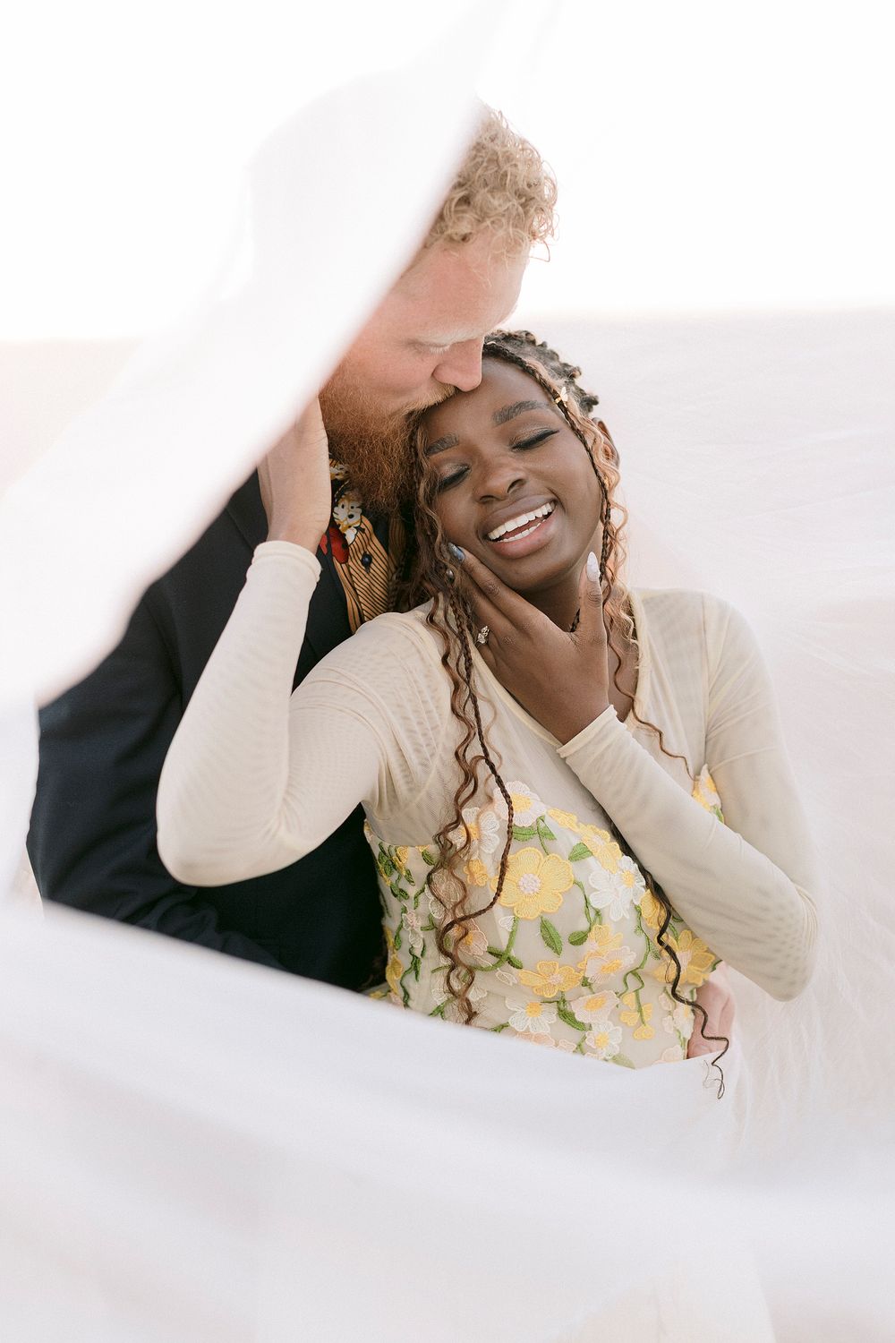 Interracial couple sharing tender moment under wedding veil, groom kissing bride's forehead as she smiles joyfully.