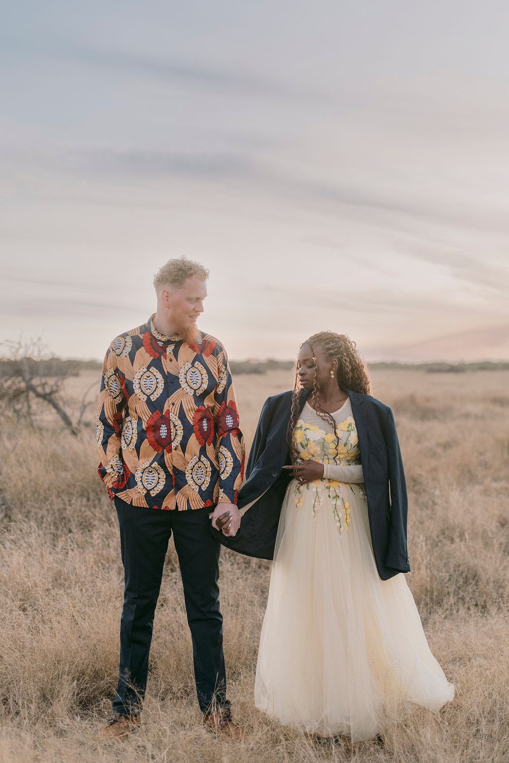 Interracial couple holding hands in a golden field, man in African print shirt, woman in white gown with blazer.