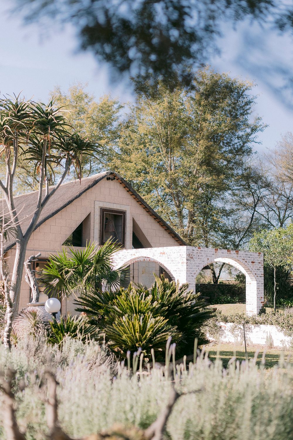 Charming white brick cottage with A-frame roof, arched gateway, and lush desert garden surrounded by tall trees.