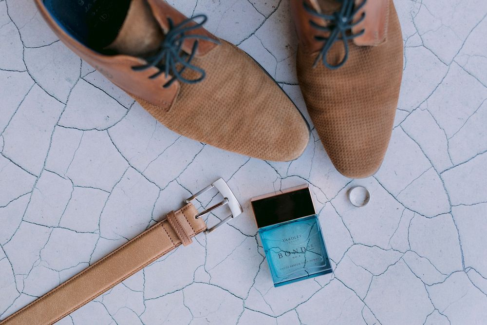 Brown suede dress shoes, leather belt, blue cologne bottle, and gold ring arranged on white cracked tile floor.