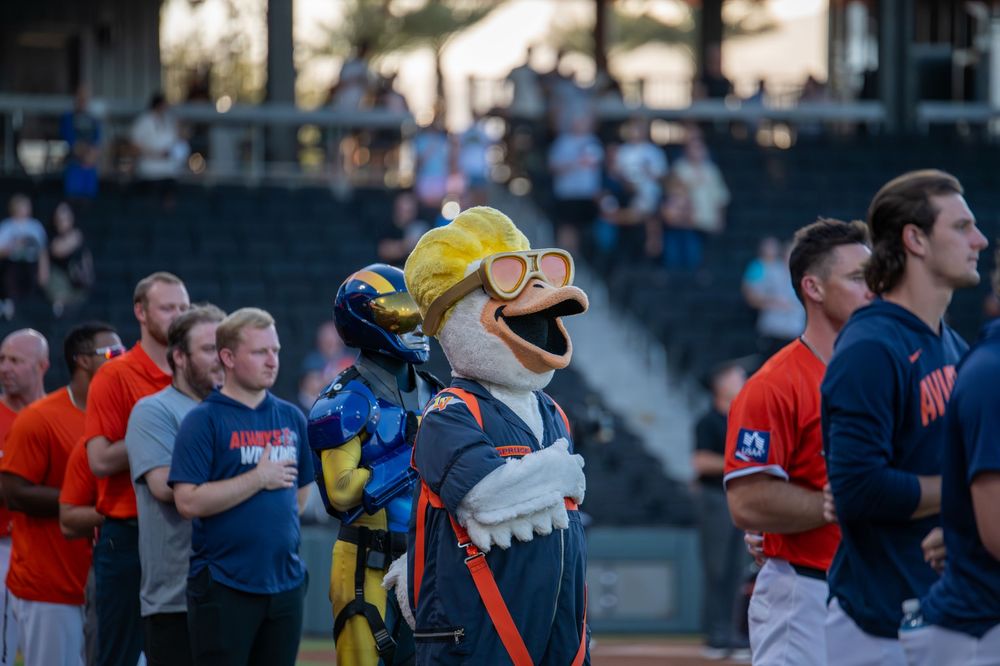 Baseball mascot dressed as a duck with yellow helmet and sunglasses stands with players on field during pregame ceremony.