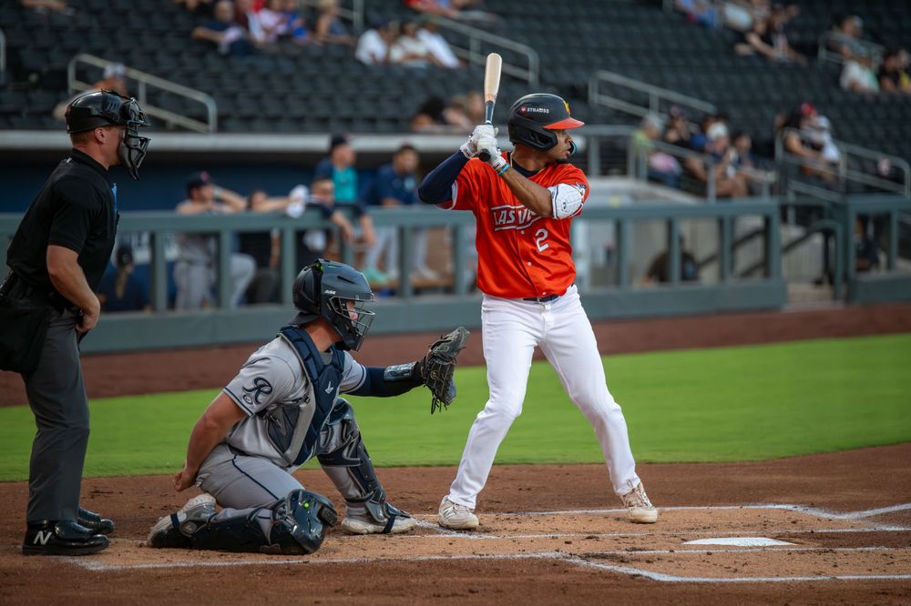 Baseball batter in orange uniform stands ready at home plate with catcher and umpire during a minor league game.