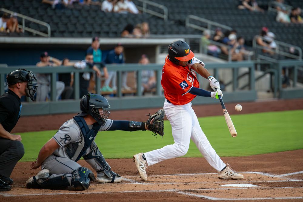 Baseball batter in orange uniform swings at pitch as catcher and umpire watch during a minor league game.