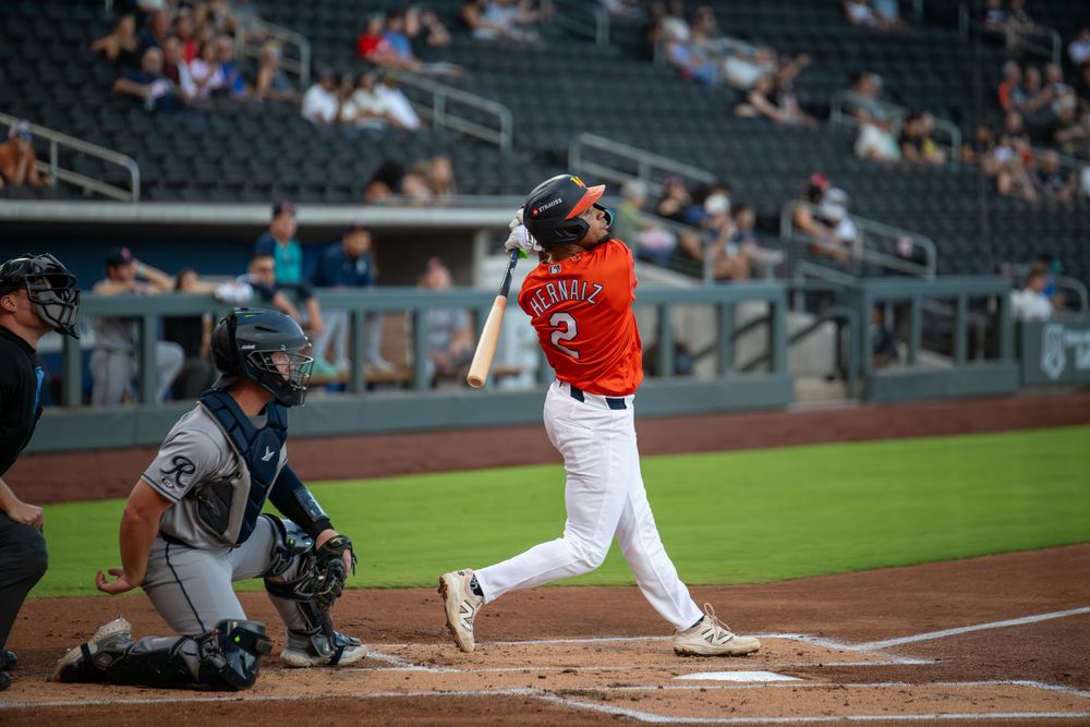 Baseball batter in orange uniform swings at pitch as catcher crouches behind home plate in stadium.