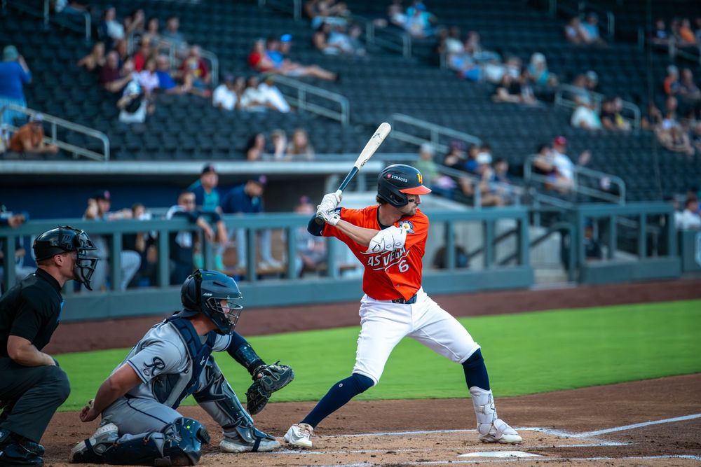 Baseball batter in orange uniform at home plate, with catcher and umpire, in a professional stadium.