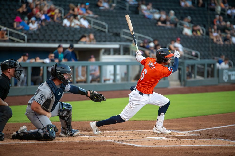 Baseball batter in orange uniform swings at pitch, with catcher and umpire behind home plate in stadium.
