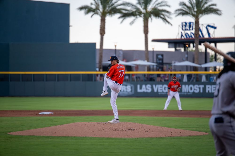 Baseball pitcher in red uniform winds up on the mound at Las Vegas ballpark with palm trees in background.