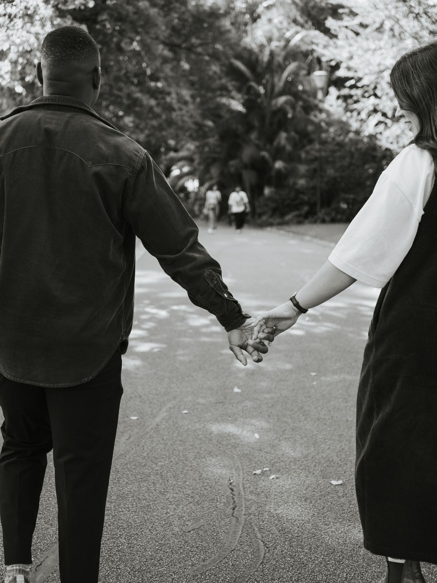Black and white photo of a couple holding hands while walking on a path surrounded by trees.