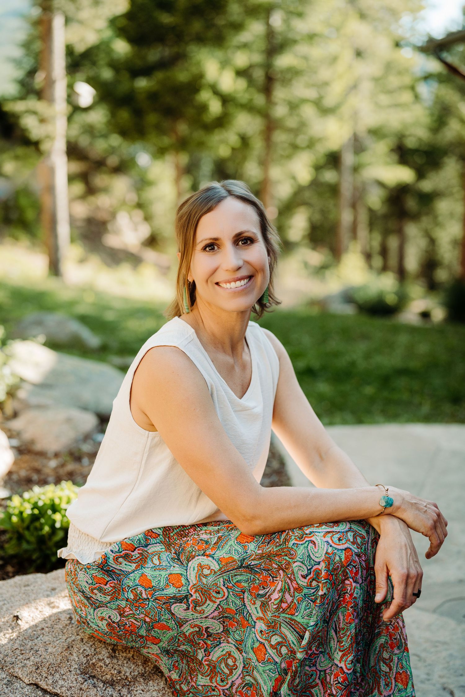 Smiling woman in floral skirt sitting on rocks outdoors in a sunny forest setting during a headshot session by McGlone Photography