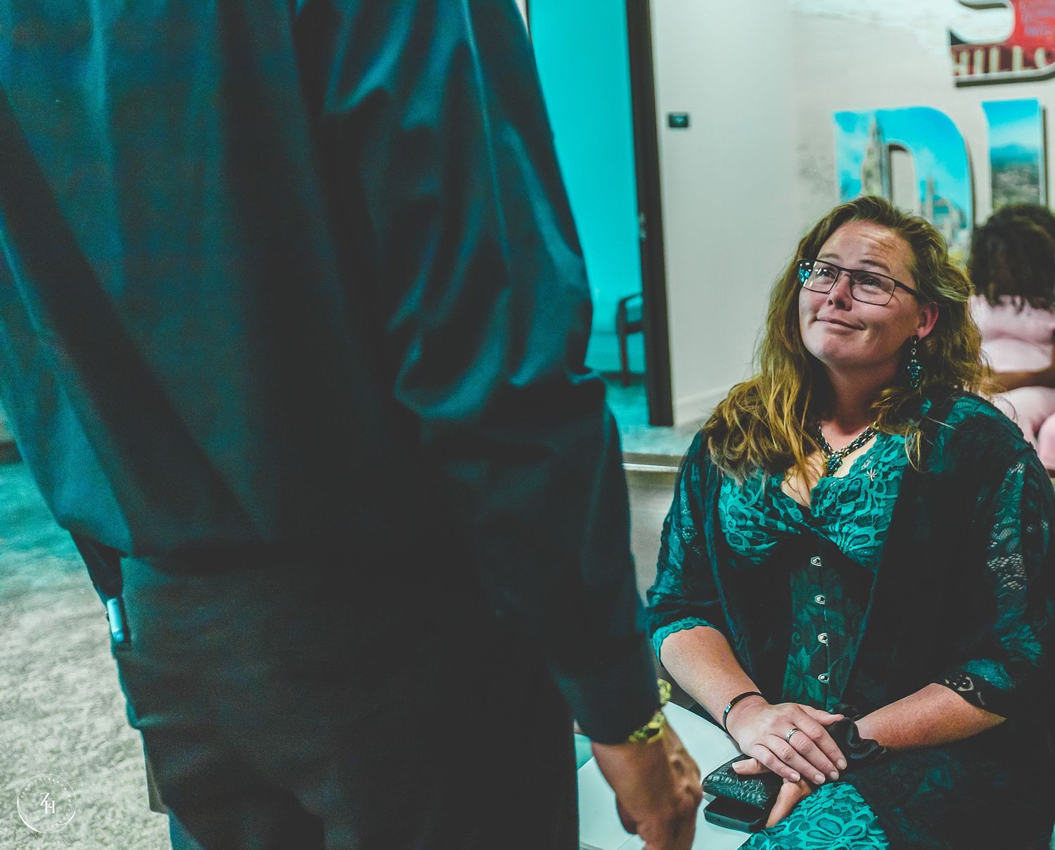 Woman with curly hair and glasses smiling warmly during an indoor event, wearing a teal patterned blouse.