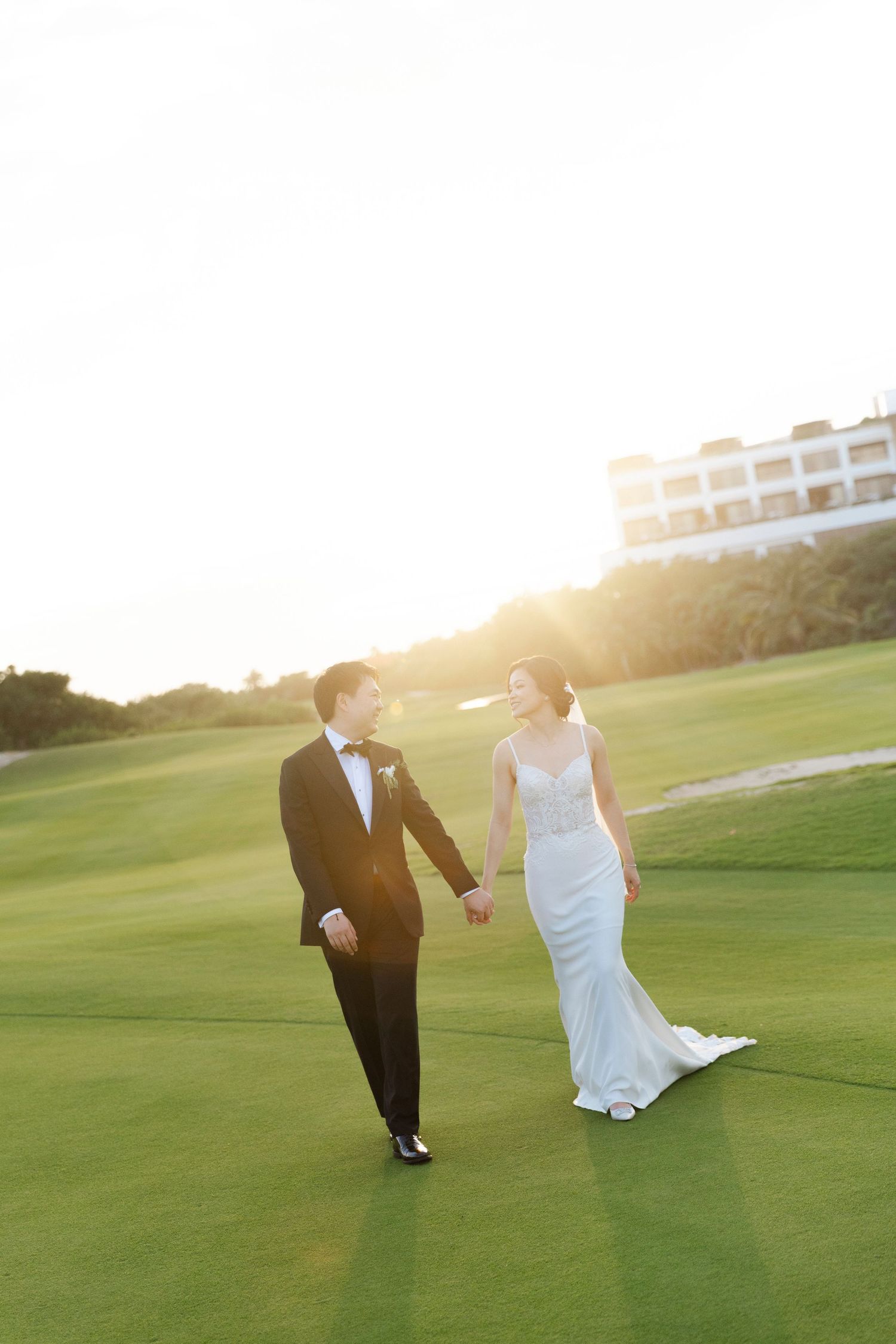Bride and groom walk hand-in-hand on a sunlit golf course at golden hour, bride in white gown, groom in black tuxedo.
