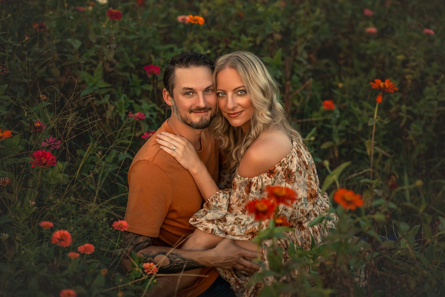 Couple sitting in a vibrant flower field with red and orange zinnias in soft focus at golden hour.