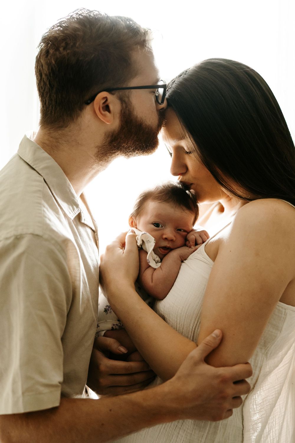 A couple tenderly kisses their newborn baby, sharing a loving family moment in soft natural light.