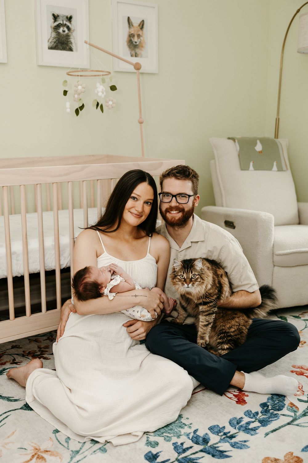 A smiling couple sits on a nursery floor holding their newborn baby and a tabby cat, with a crib and animal art behind them.