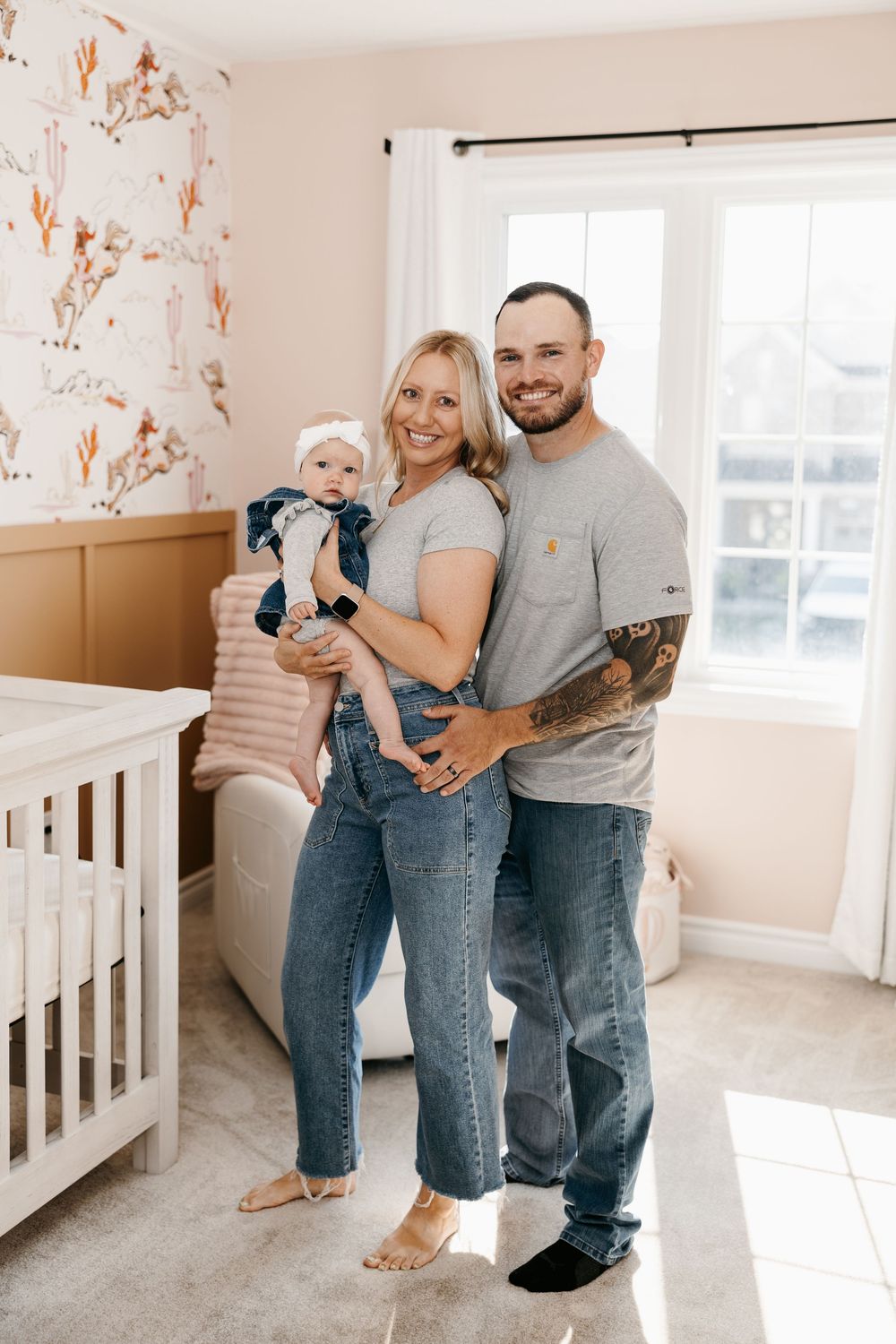 A smiling couple in matching gray tees and jeans holds their baby girl in a softly decorated nursery.