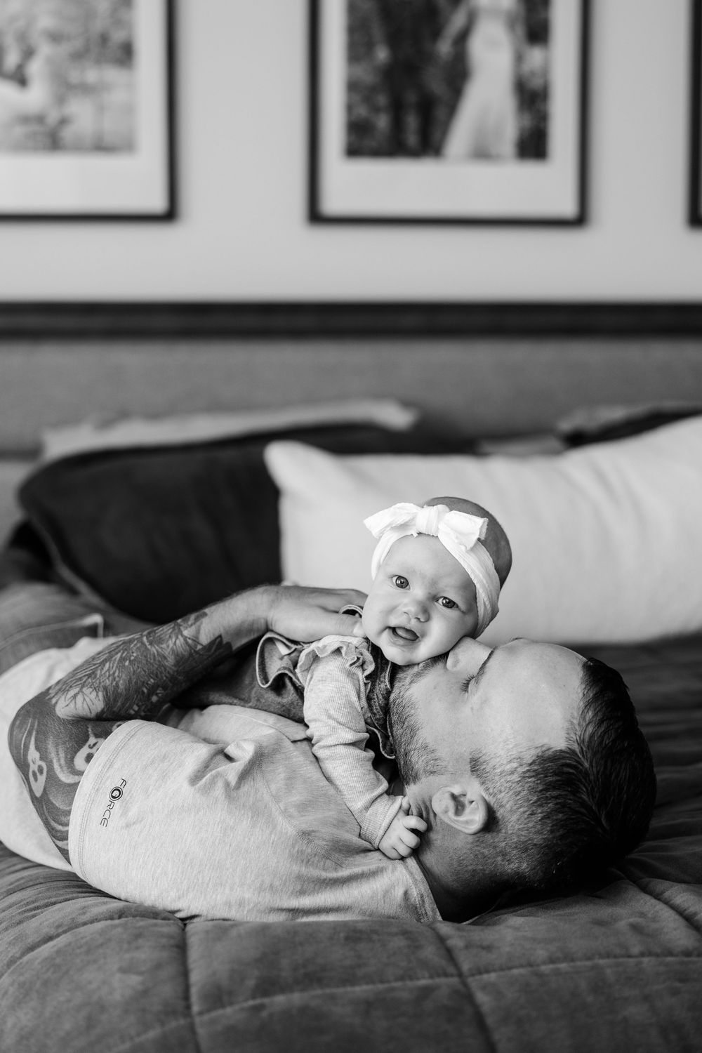 Black and white photo of tattooed father lying on bed with smiling baby girl wearing a white bow headband.
