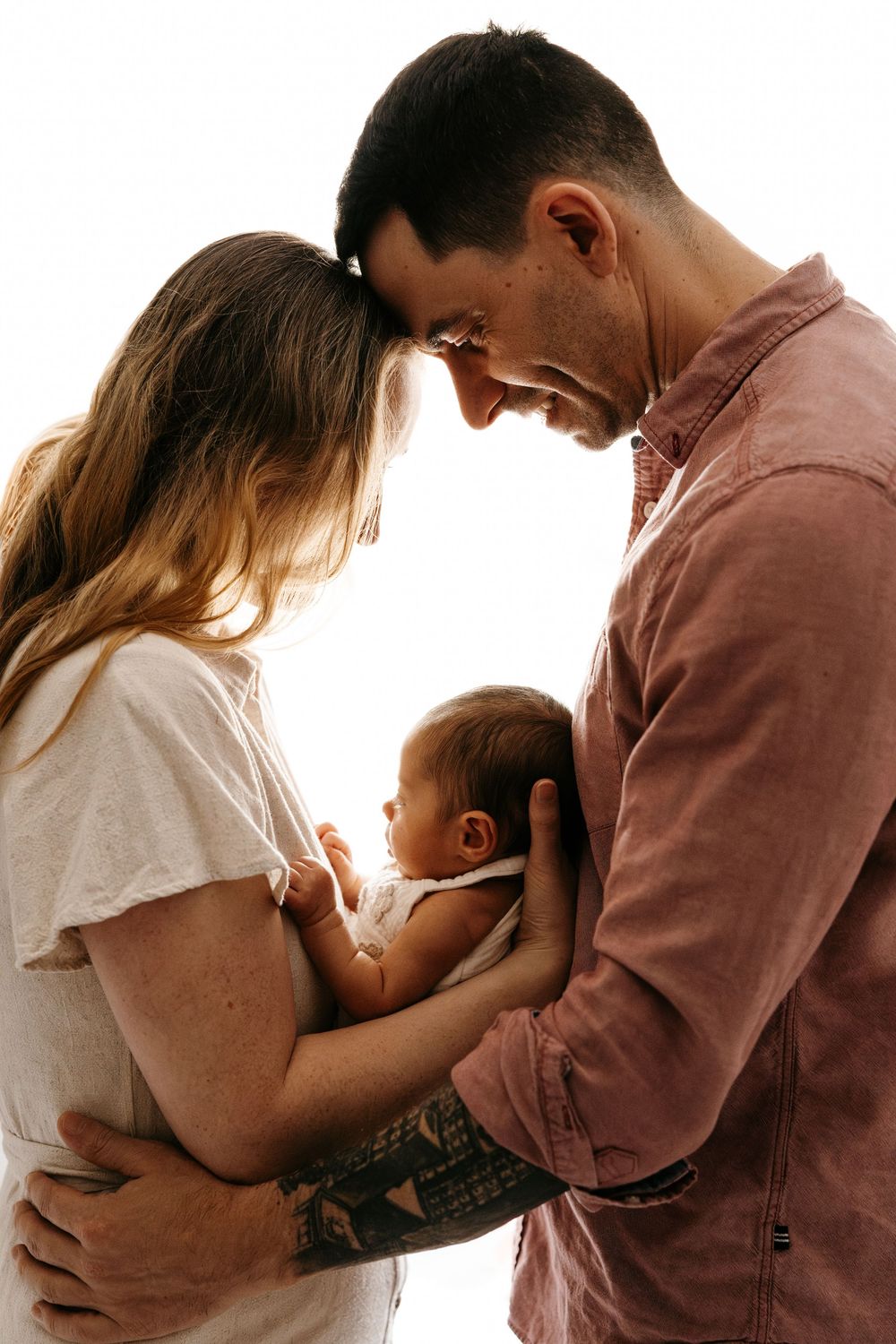 Young parents smiling down at their newborn baby, backlit by soft white light in an intimate family portrait.