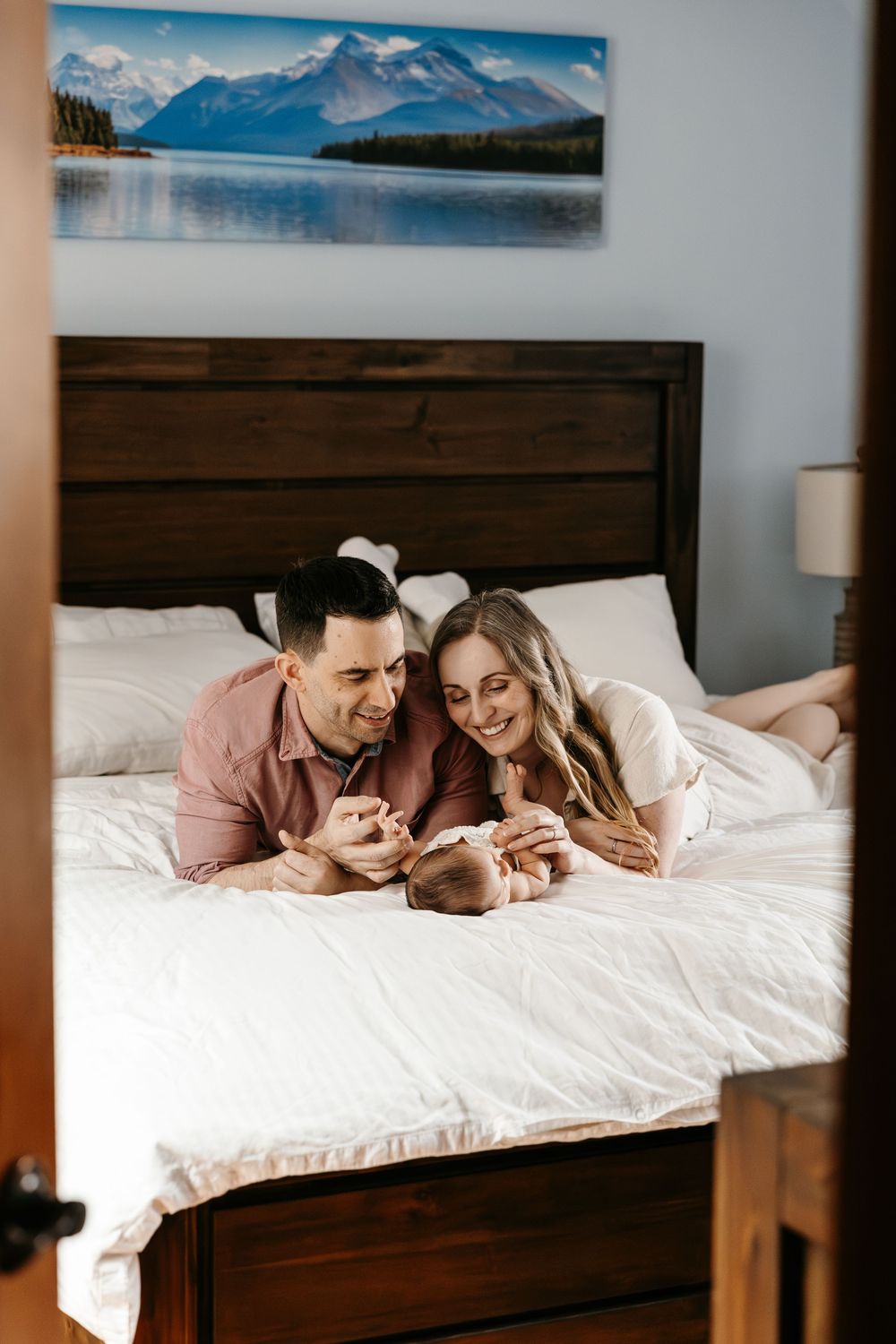 A happy couple lies on a white bed smiling at their newborn baby in a cozy bedroom with mountain artwork.