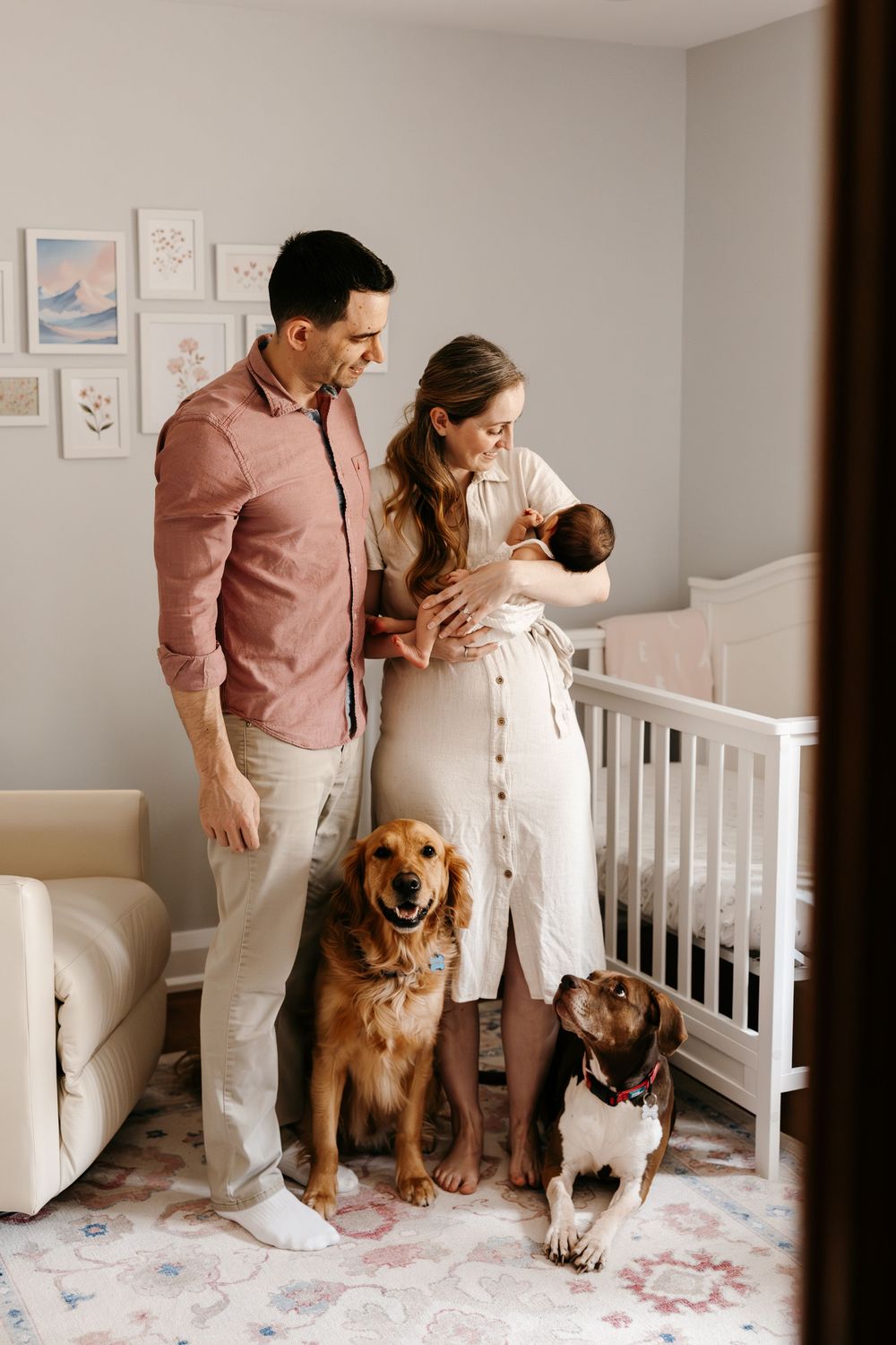 A young family with a newborn baby and two dogs poses in a nursery beside a white crib.