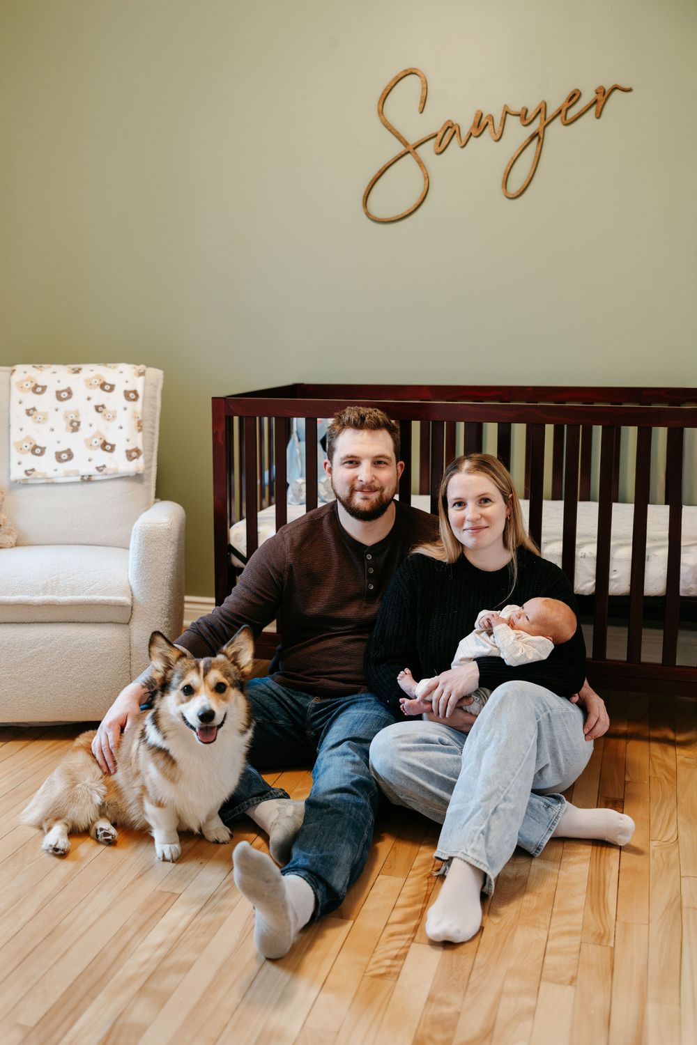 Young couple sitting on nursery floor with corgi dog and newborn baby in front of crib with Sawyer name sign.
