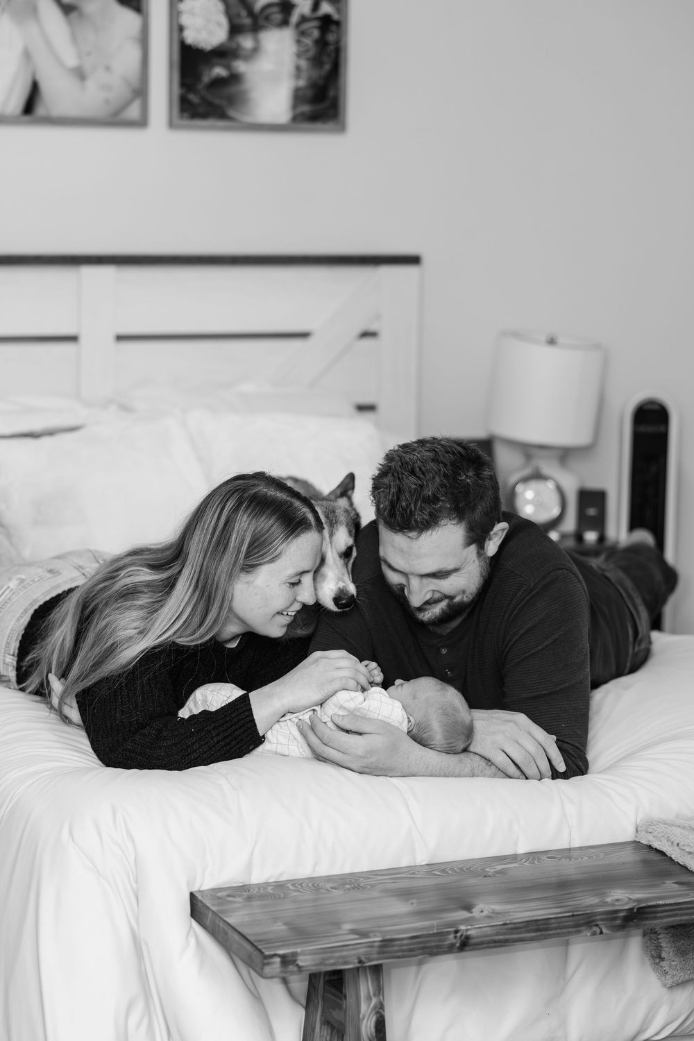 Black and white photo of parents lying on bed with newborn baby and dog, sharing a tender family moment.