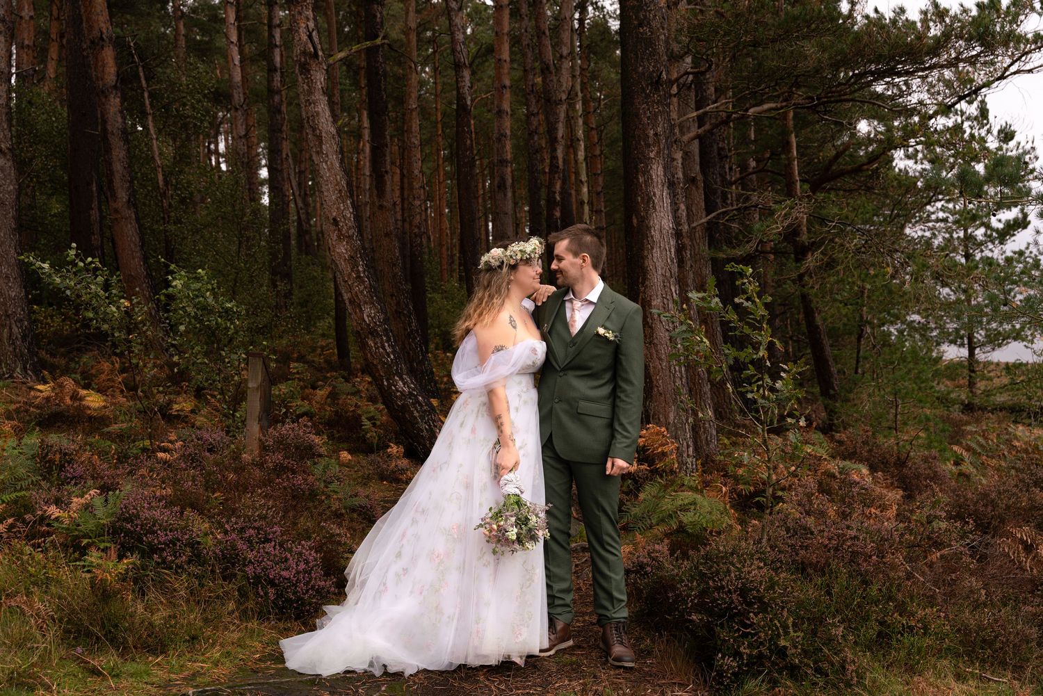 A bride and groom share a romantic moment in a pine forest, bride wearing a floral crown and white gown.