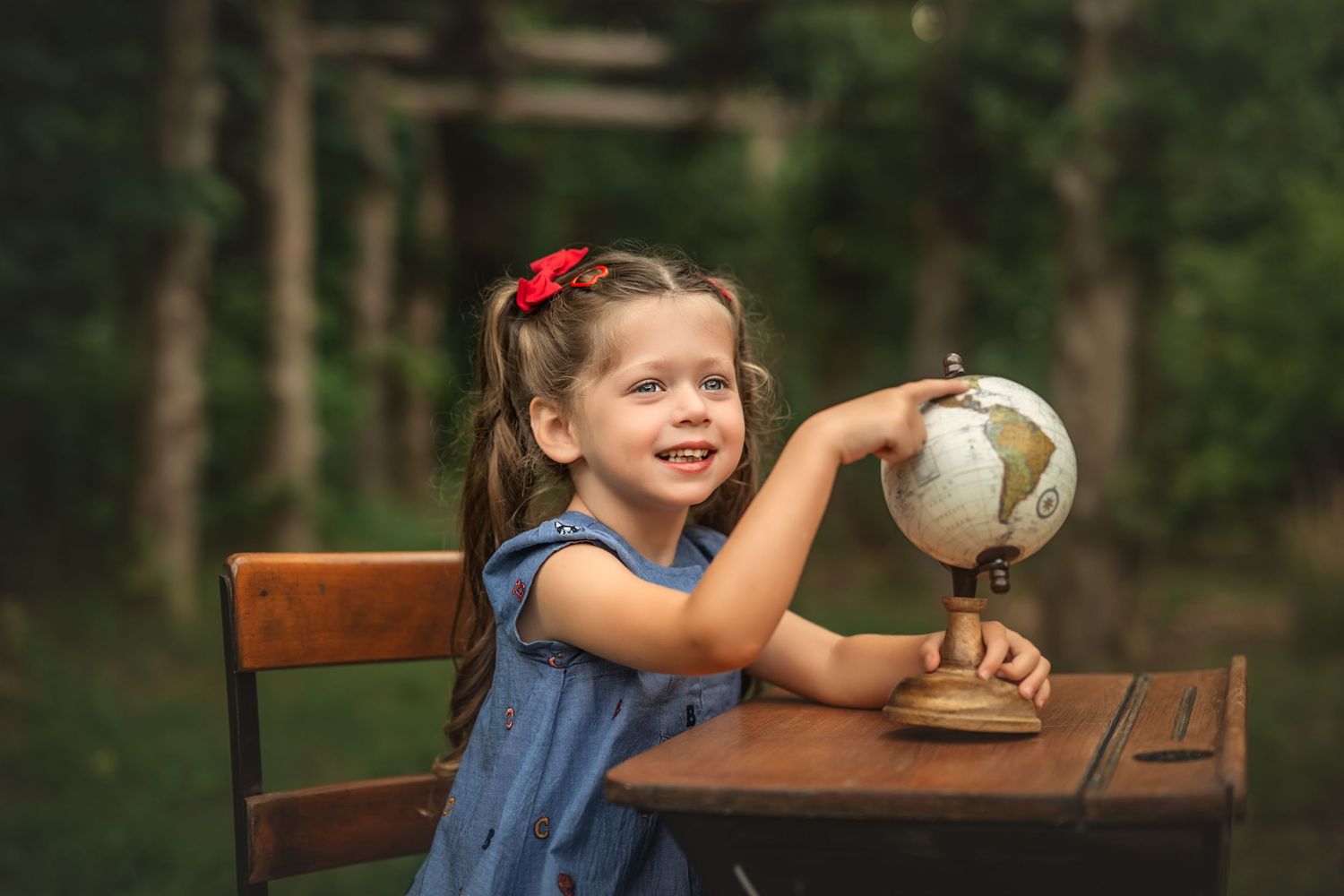 A smiling little girl in a blue dress pointing at a vintage globe on a wooden desk outdoors.
