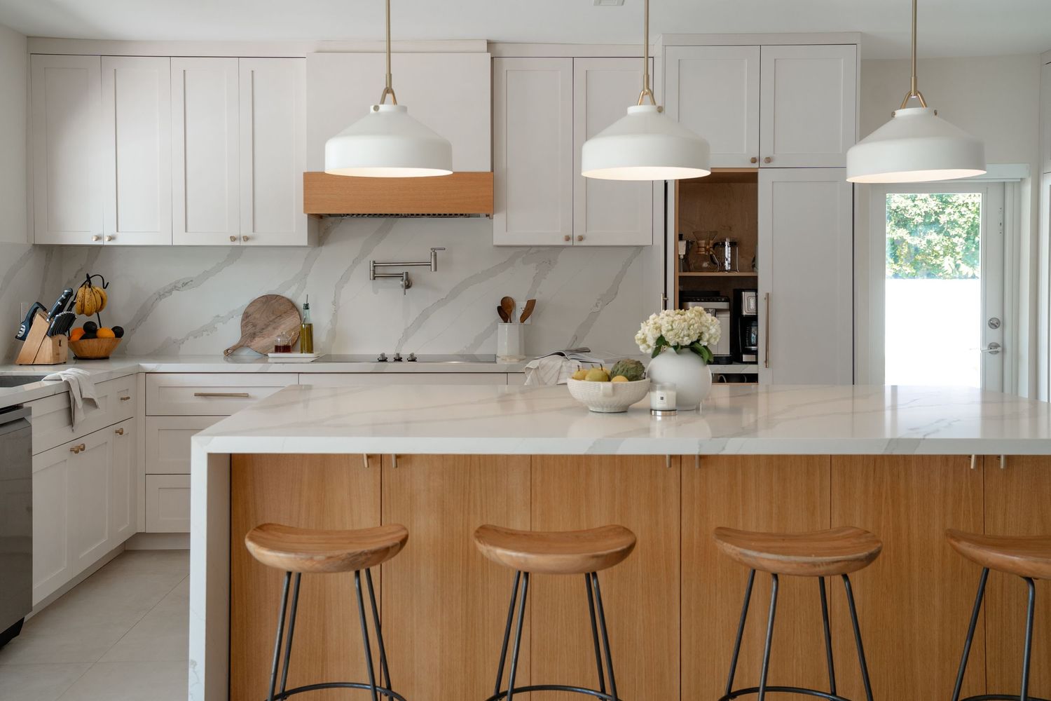 Modern white kitchen with marble backsplash, wood island, white pendant lights, and bar stools.
