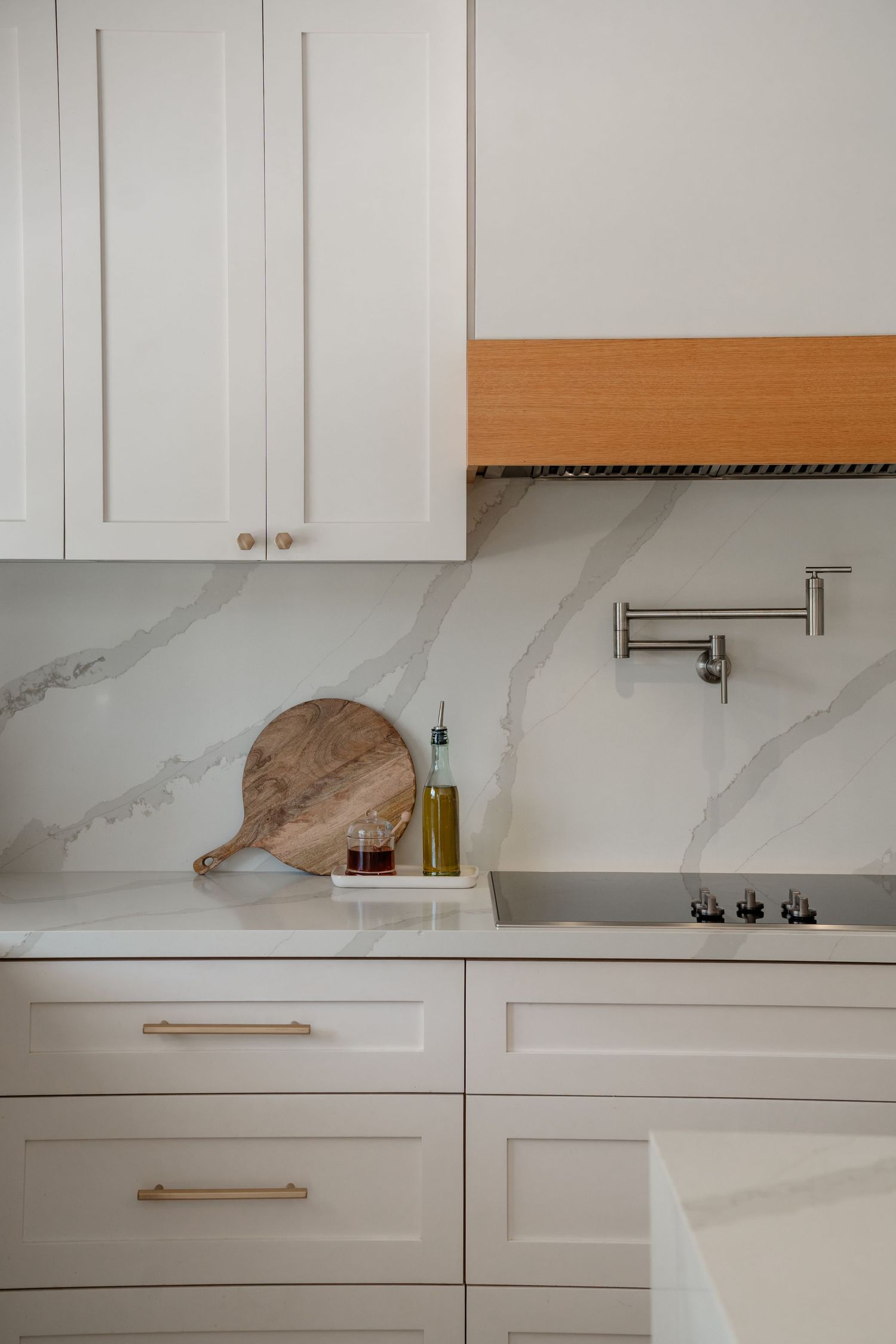 White shaker kitchen cabinets with brass hardware, marble backsplash, pot filler, and wooden cutting board on quartz countertop.