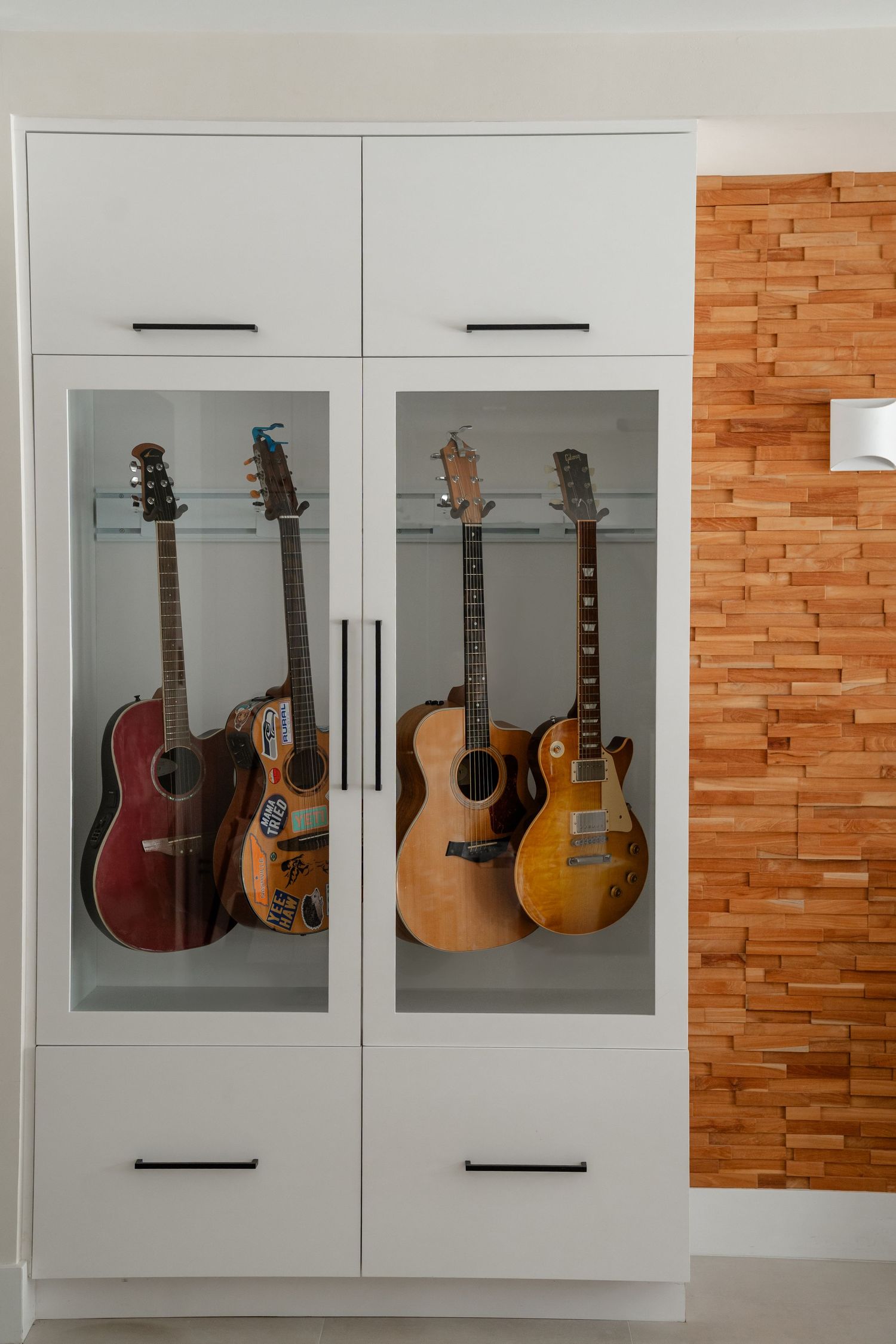 White cabinet with glass doors displaying a collection of acoustic and electric guitars beside a wood panel wall.
