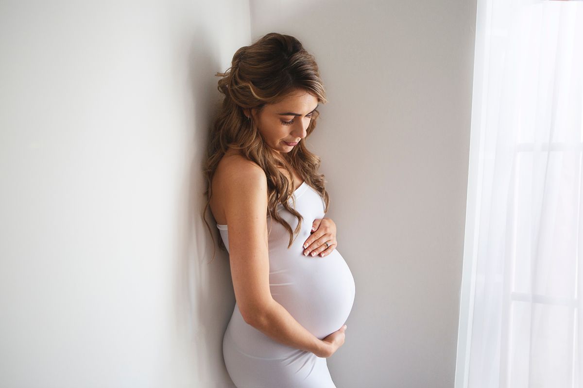 Pregnant woman in white dress cradling her baby bump while leaning against a wall near a window. during a maternity photoshoot in Kent