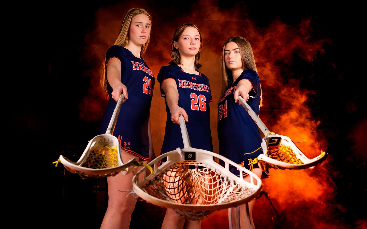 Three Hershey lacrosse seniors in a 2026 portrait with orange smoke, by Deanna Fox, SportsFox Photography based in Hershey PA