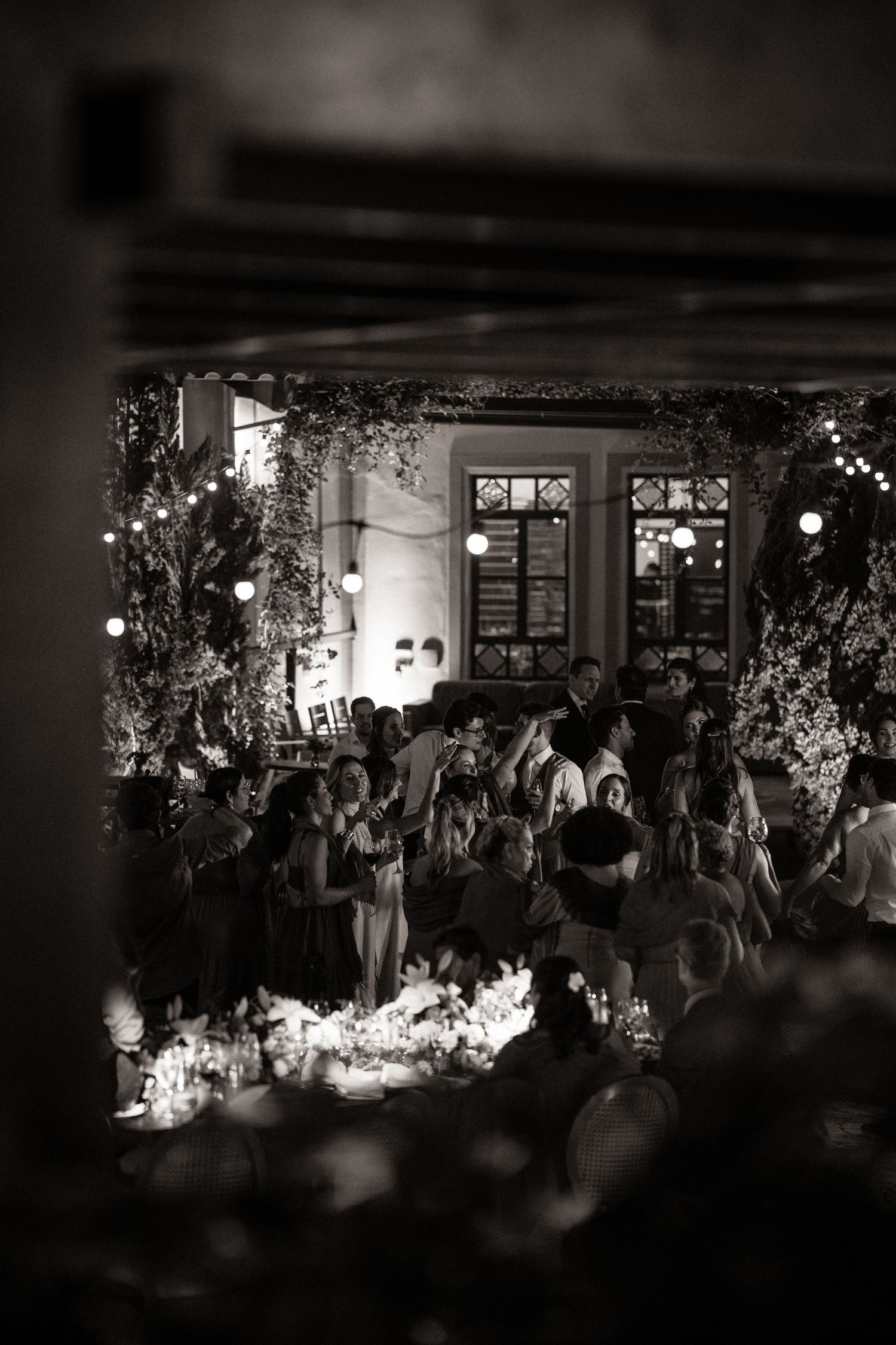 Black and white photo of elegant evening wedding reception with guests gathered under string lights and lush floral arrangements.