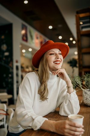 A stylish woman in a red hat and white sweater enjoys coffee at a cozy cafe.