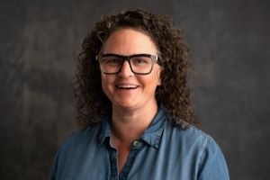 Professional headshot of someone with curly hair wearing glasses against a dark background.