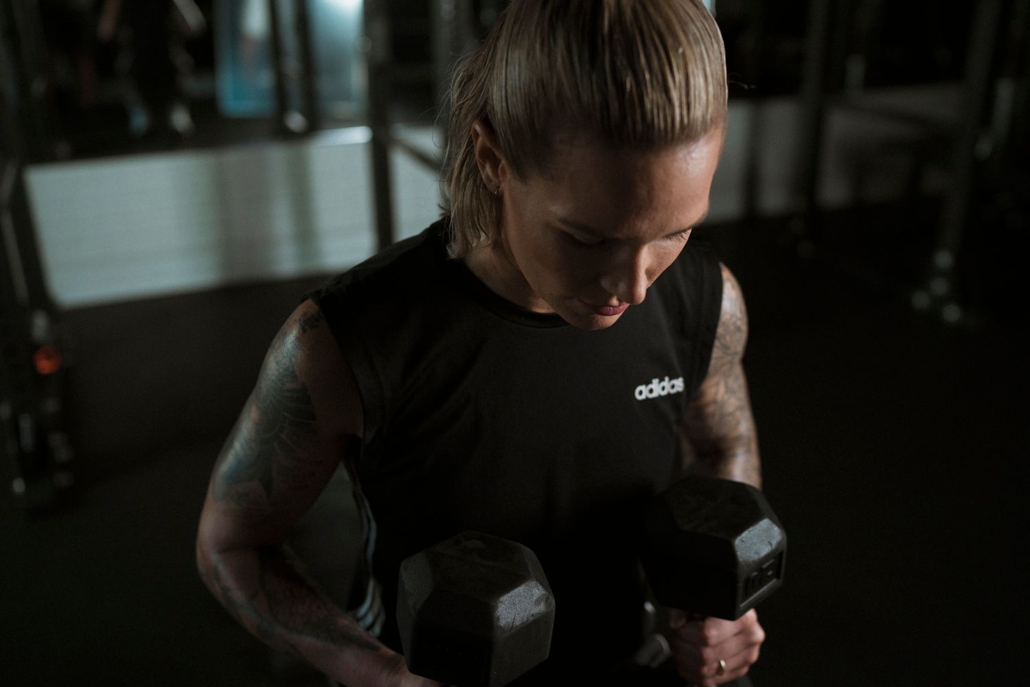 Athletic person in black workout gear lifting dumbbells in a dimly lit gym setting.