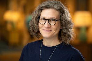 Professional portrait of someone with wavy grey hair wearing glasses, a navy blue top and necklace against a blurred background.