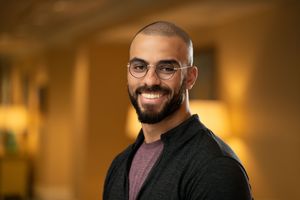Professional headshot of a smiling person with glasses and beard wearing a black cardigan over a mauve shirt in warm lighting.