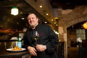 A chef in black uniform stands with wine glass in a rustic restaurant interior.