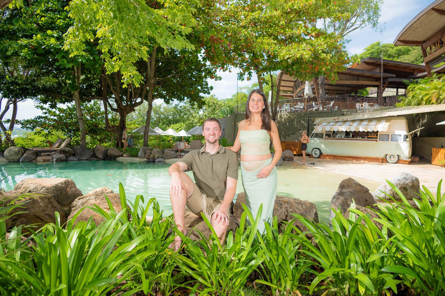 Couple posing by a tropical resort pool surrounded by lush greenery and rocks on a sunny day.