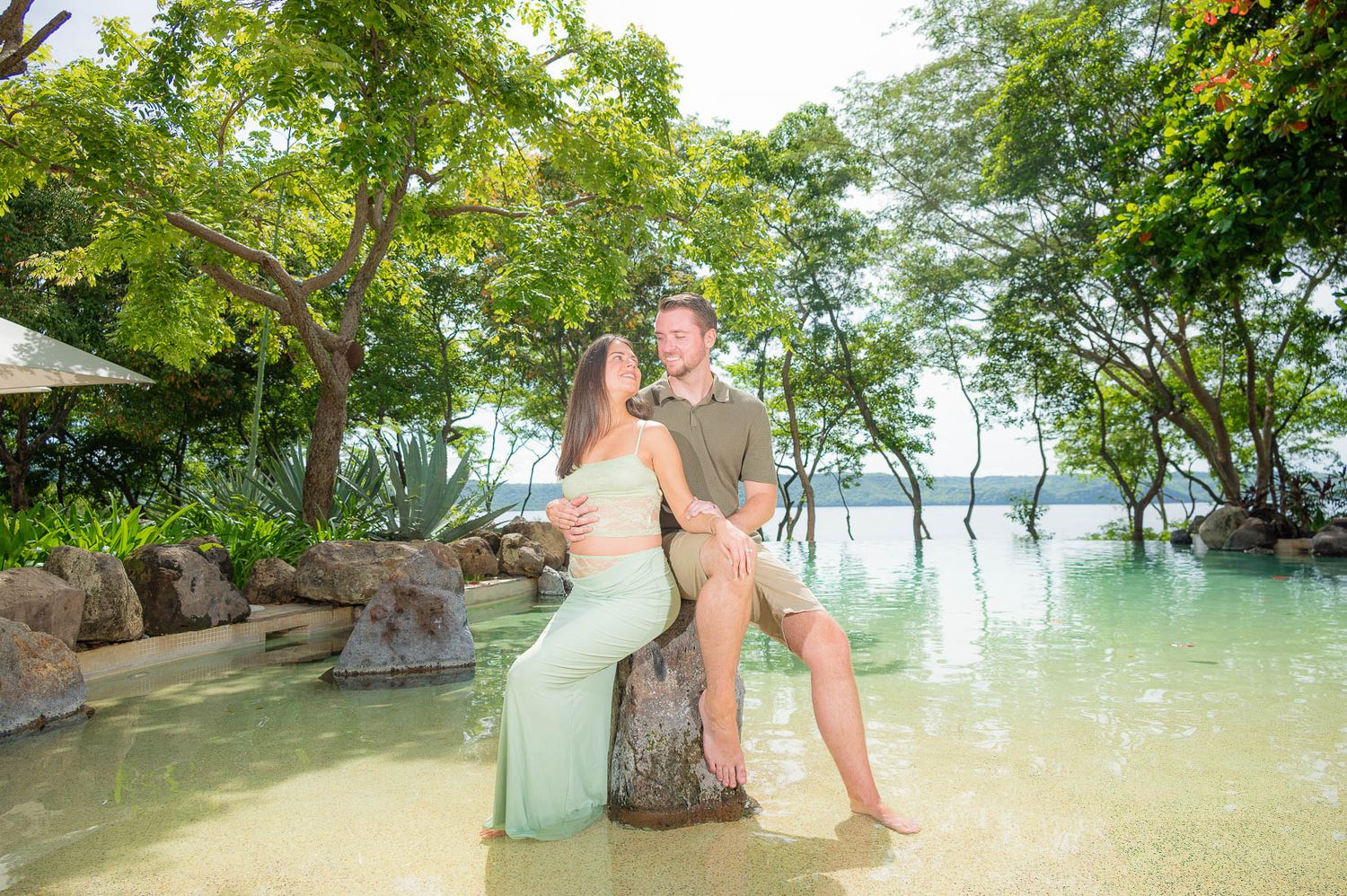 Couple posing romantically by a tropical resort pool with lush green trees and calm ocean waters in the background.