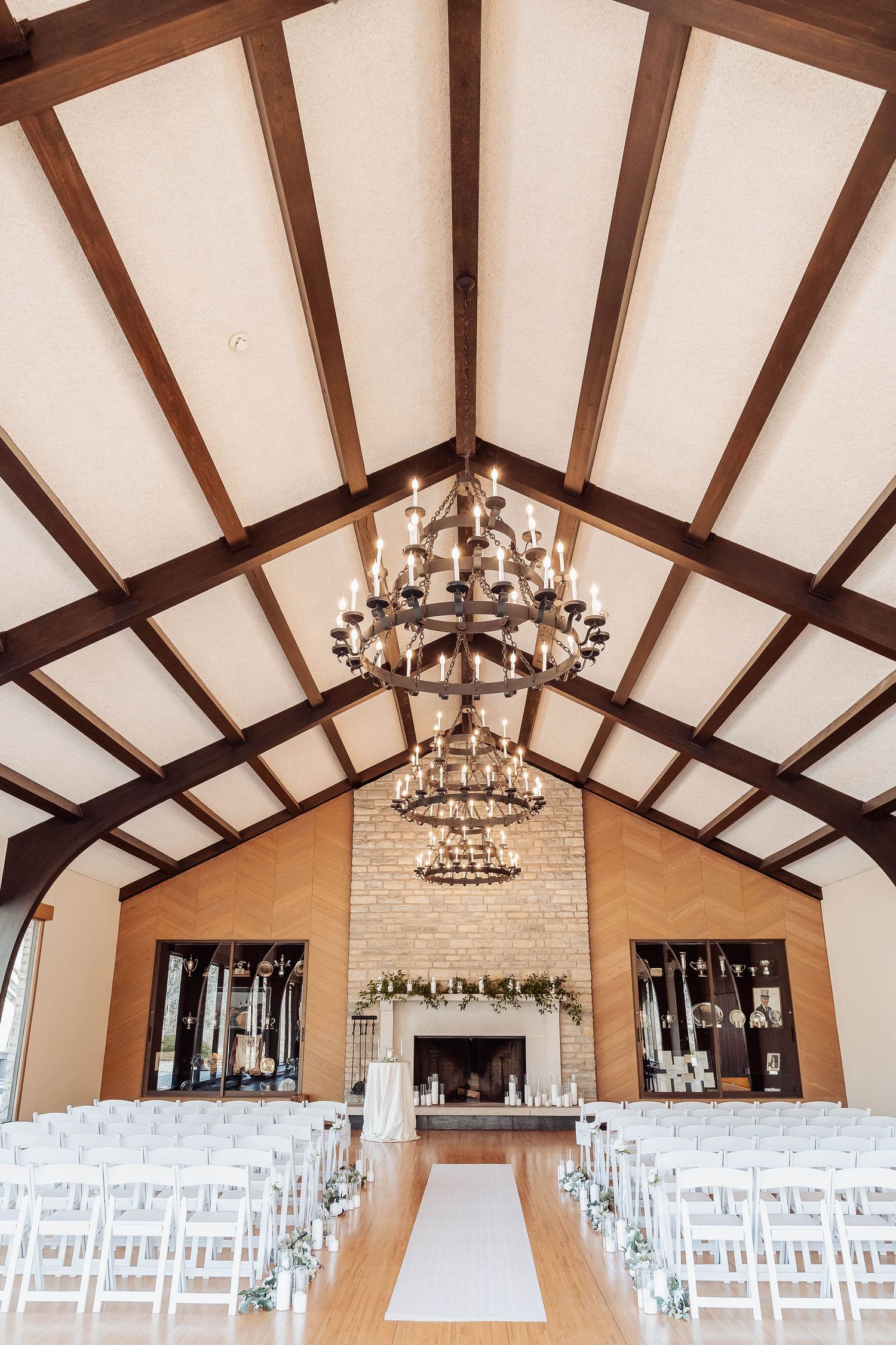 Elegant wedding venue with vaulted wood beam ceiling, chandelier, white chairs, and brick fireplace altar.