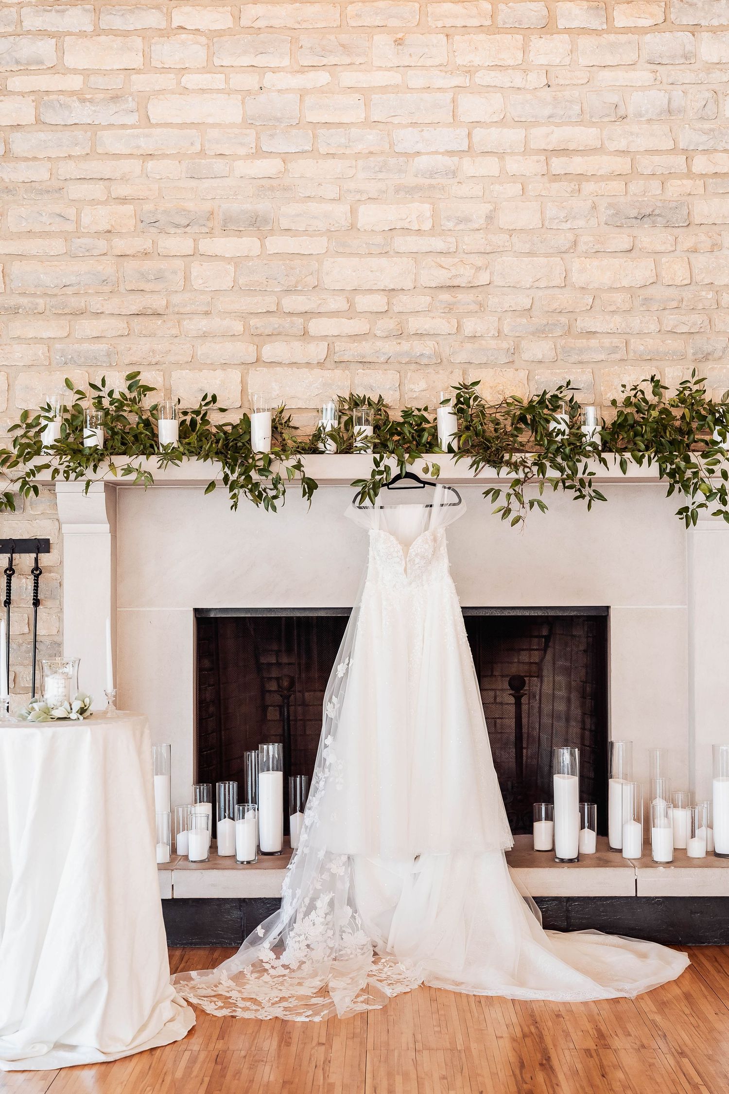 Elegant wedding dress hanging over a white fireplace adorned with greenery and candles in a rustic venue.