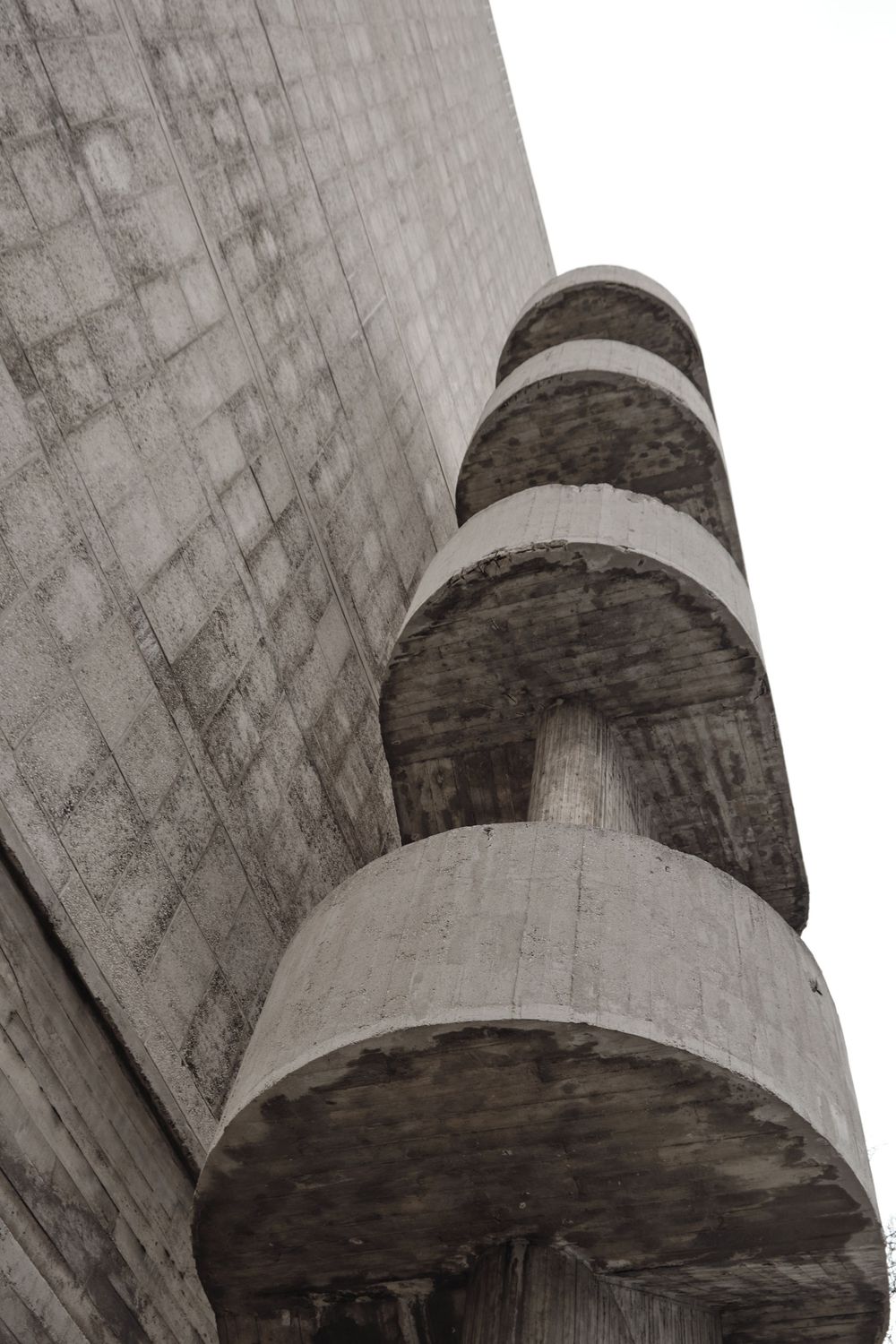 Low-angle view of a brutalist concrete spiral staircase with stacked circular landings curving up the side of a textured concrete wall.