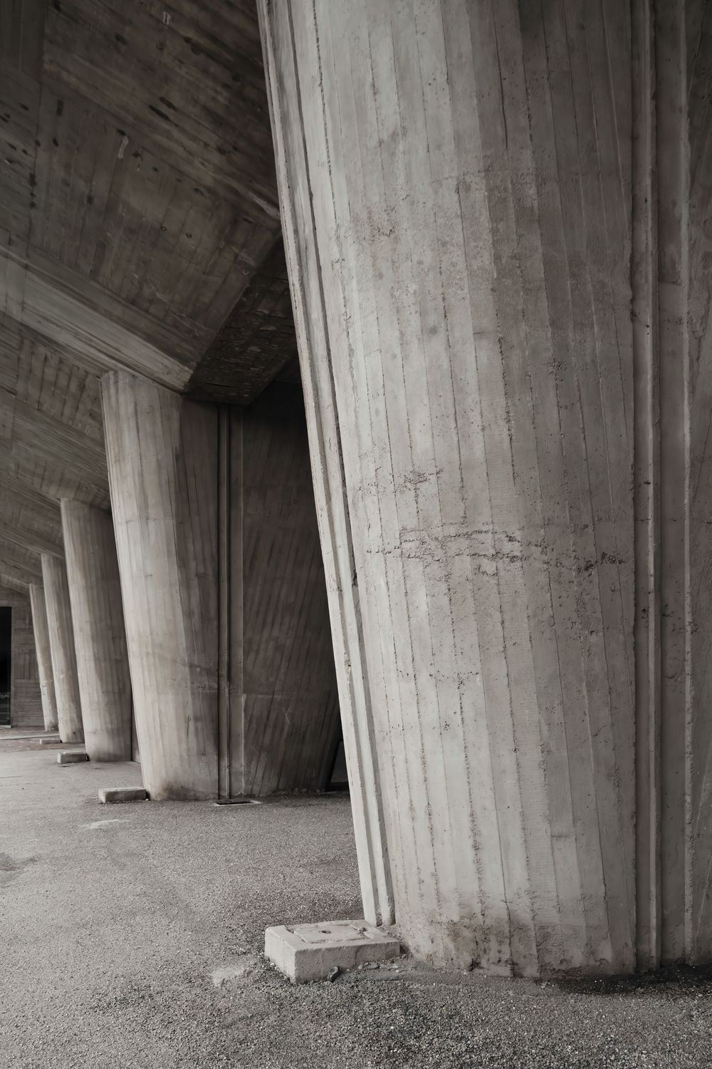 Massive board-formed concrete pillars lining a shadowed brutalist colonnade with a rough gravel floor and textured wood-grain surfaces.
