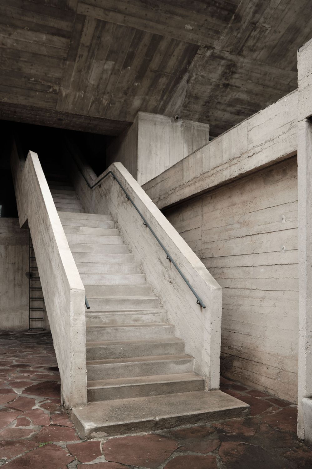 Brutalist concrete staircase with metal handrail rises beside board-formed concrete walls over a rough stone-paved floor.
