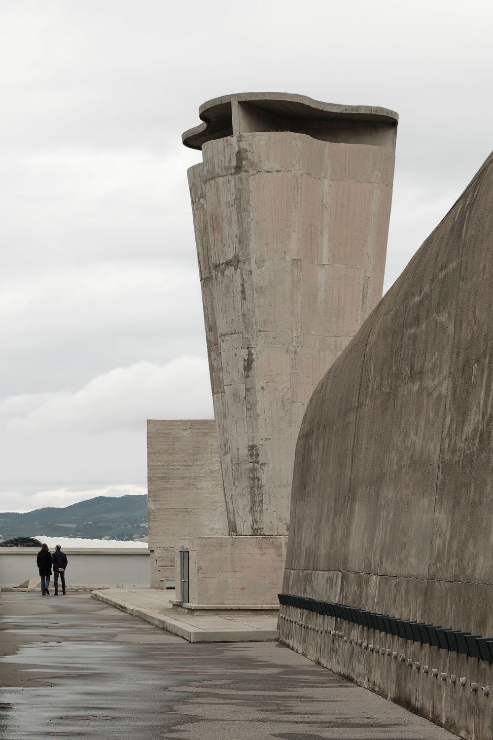 Two people walk along the weathered concrete rooftop terrace of a brutalist building, with a sculptural ventilation tower and hills beyond.